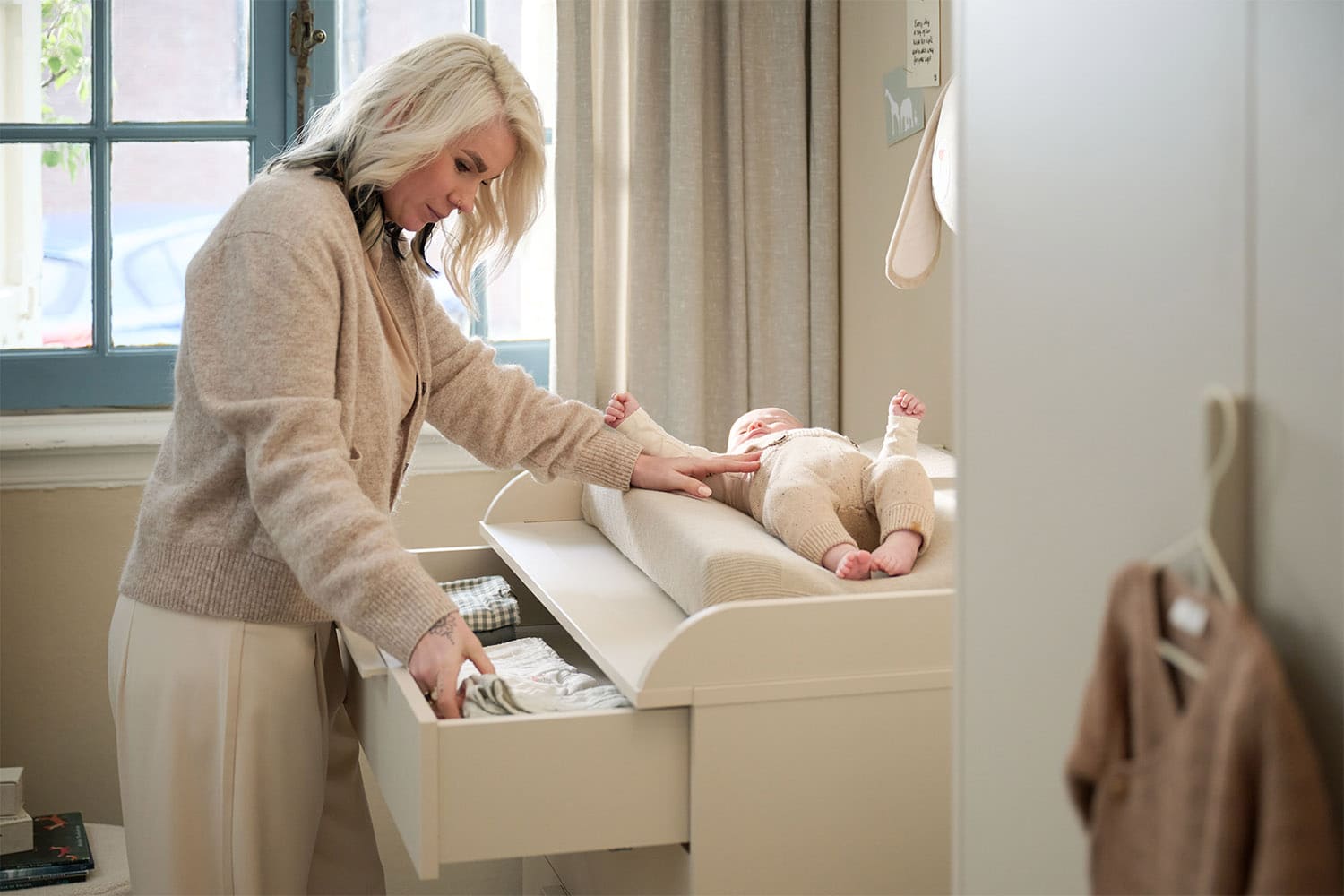 Une femme change un bébé sur une table à langer beige, tout en prenant des vêtements dans un tiroir ouvert dans une chambre lumineuse.